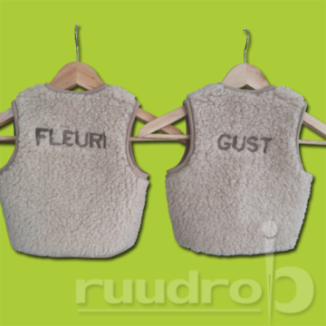 Twee wollen beige baby bodywarmertjes met de namen Fleuri en Gust erop geborduurd